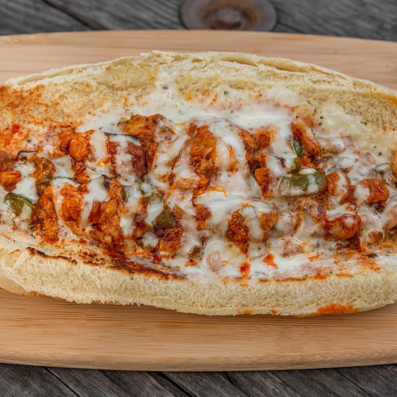 A close-up of a chicken cheesesteak sandwich with melted white cheese and red sauce on a wooden board.